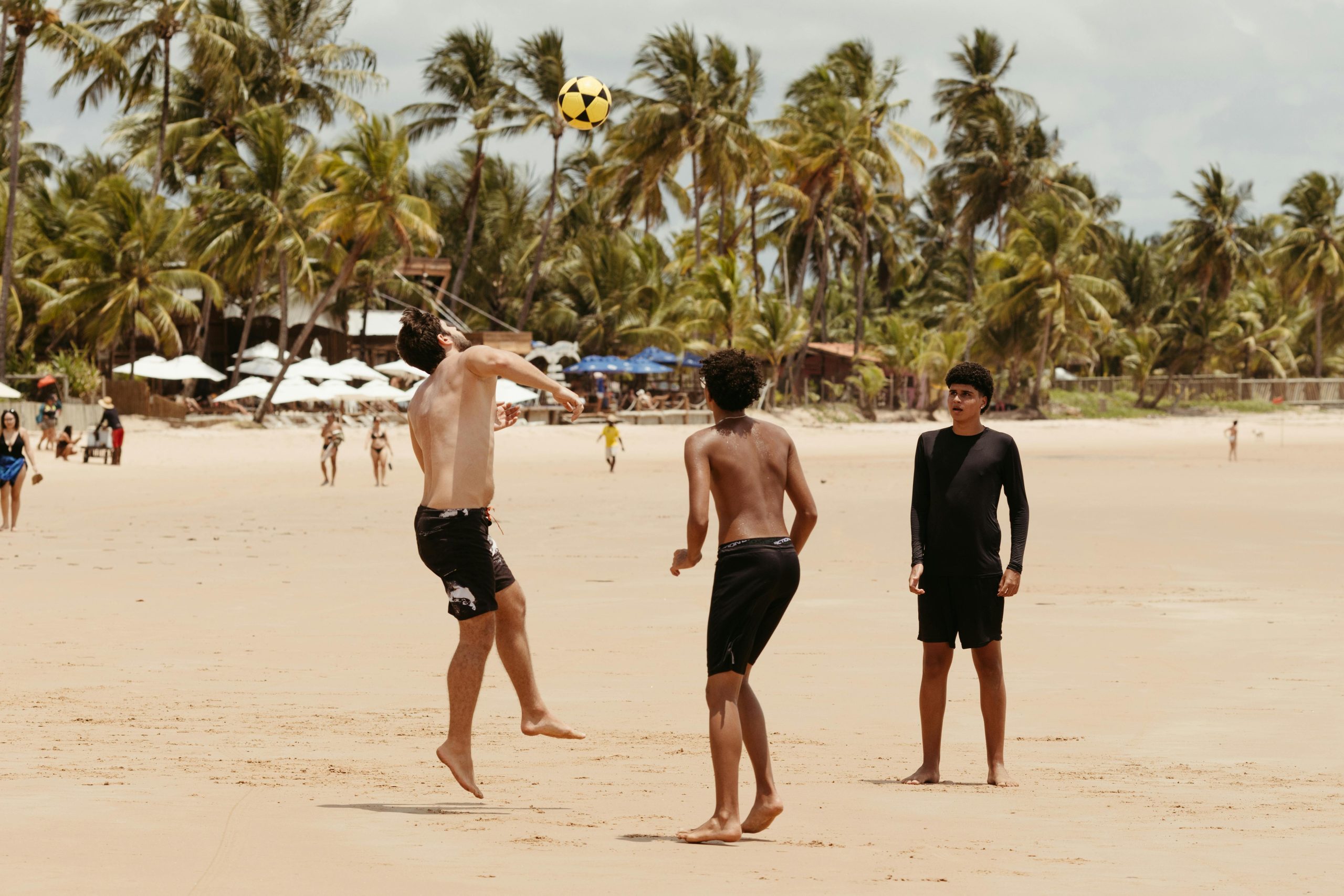 Group of teenagers playing beach soccer under palm trees on a sunny day.